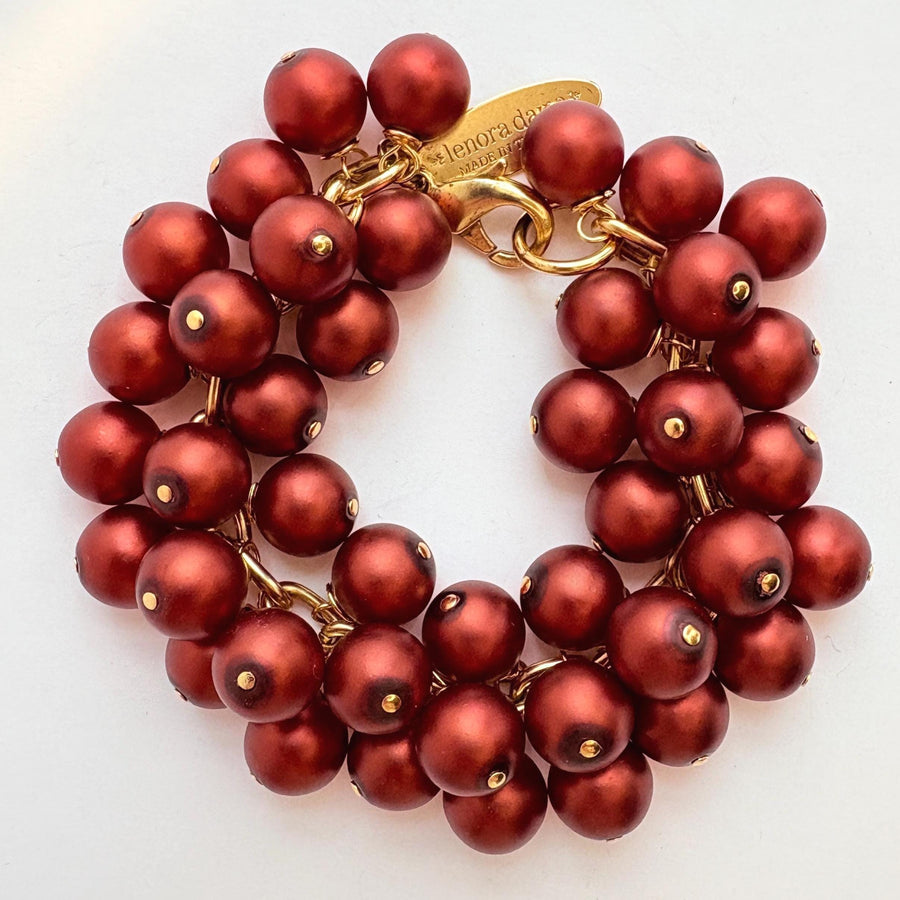 A gold chain bracelet with multiple strands of matte dark red faux pearls. The bracelet has a small tag that reads &#39;Lenora Dame&#39; and &#39;Made in the USA.&#39;