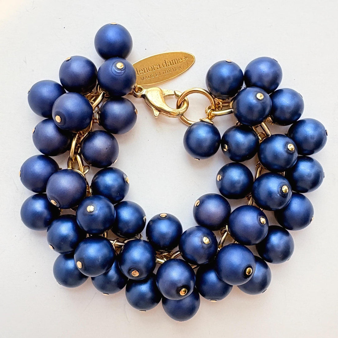 A gold chain bracelet with multiple strands of matte navy blue faux pearls. The bracelet has a small tag that reads &