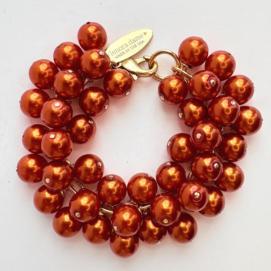 A gold chain bracelet with multiple strands of shiny rust orange faux pearls. The bracelet has a small tag that reads &#39;Lenora Dame&#39; and &#39;Made in the USA.&#39;