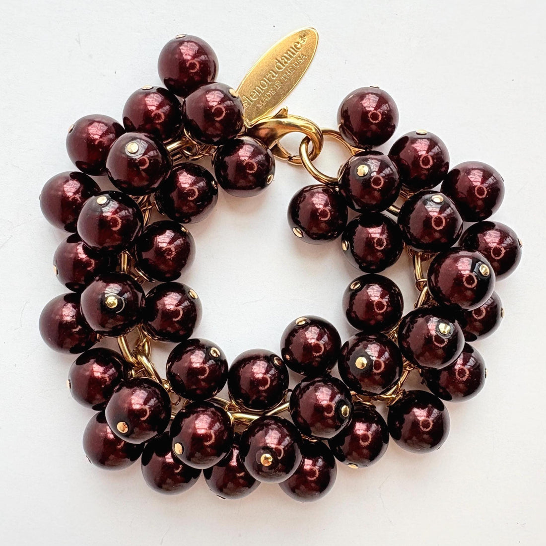 A gold chain bracelet with multiple strands of shiny dark burgundy faux pearls. The bracelet has a small tag that reads &
