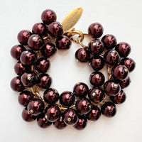 A gold chain bracelet with multiple strands of shiny dark burgundy faux pearls. The bracelet has a small tag that reads &#39;Lenora Dame&#39; and &#39;Made in the USA.&#39;