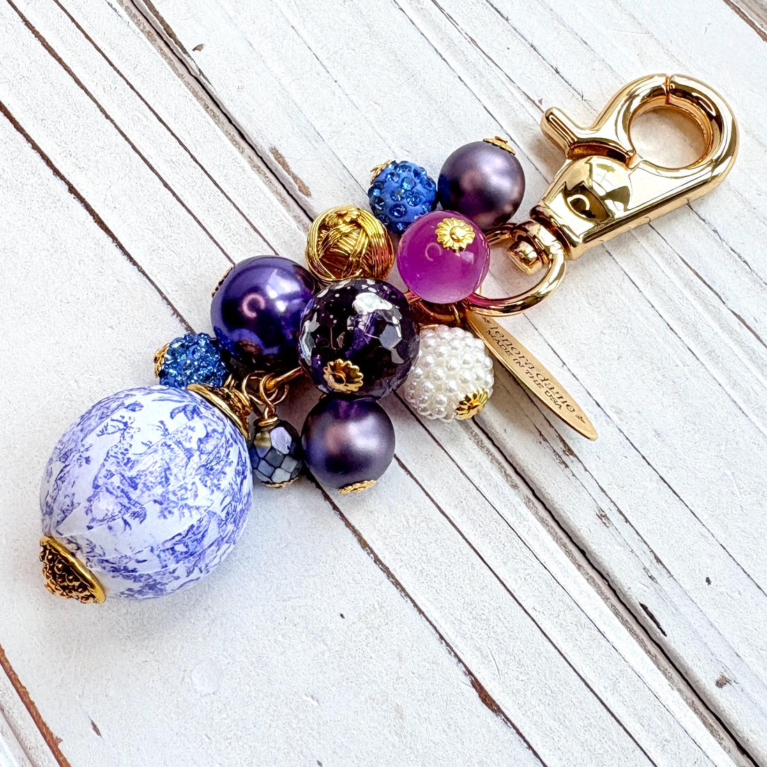 Beaded bag charm with a mix of bead styles in shades of purple, blue, gold and pearl. One larger round bead in a purple and white toile print hangs at the bottom and a gold trigger clasp on top of all of the beads. Brass oval hang tag near the trigger clasp that says &quot;Lenora Dame Handmade in USA&quot;. Lenora Dame bag charm.