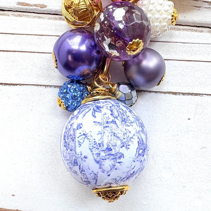 Beaded bag charm with a mix of bead styles in shades of purple, blue, gold and pearl. One larger round bead in a purple and white toile print hangs at the bottom and a gold trigger clasp on top of all of the beads. Lenora Dame bag charm.