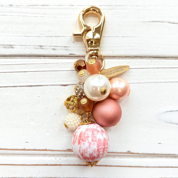 Beaded bag charm with a mix of bead styles in shades of pink, peach, gold and pearl. One larger round bead in a pink and white toile print hangs at the bottom and a gold trigger clasp on top of all of the beads. Brass oval hang tag near the trigger clasp that says &quot;Lenora Dame Handmade in USA&quot;. Lenora Dame bag charm.
