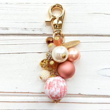 Beaded bag charm with a mix of bead styles in shades of pink, peach, gold and pearl. One larger round bead in a pink and white toile print hangs at the bottom and a gold trigger clasp on top of all of the beads. Brass oval hang tag near the trigger clasp that says &quot;Lenora Dame Handmade in USA&quot;. Lenora Dame bag charm.