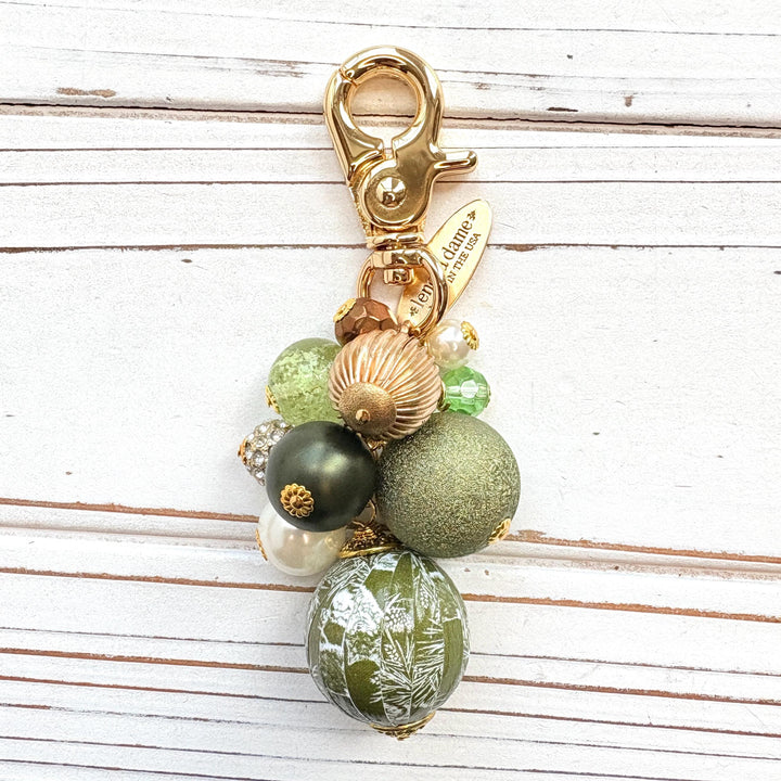 Beaded bag charm with a mix of bead styles in shades of green, gold, and pearl. One larger round bead in a army green and white toile print hangs at the bottom and a gold trigger clasp on top of all of the beads. Brass oval hang tag near the trigger clasp that says &quot;Lenora Dame Handmade in USA&quot;. Lenora Dame bag charm.