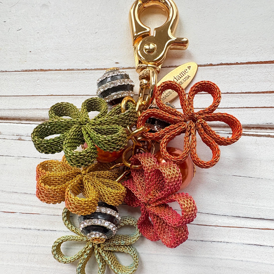 Floral bag charm with four mesh metal flowers in fall colors of green, orange and yellow, black and white striped beads with rhinestones and pumpkin orange pearls. Gold trigger clasp at the top with a gold oval charm that says Lenora Dame Made in USA.
