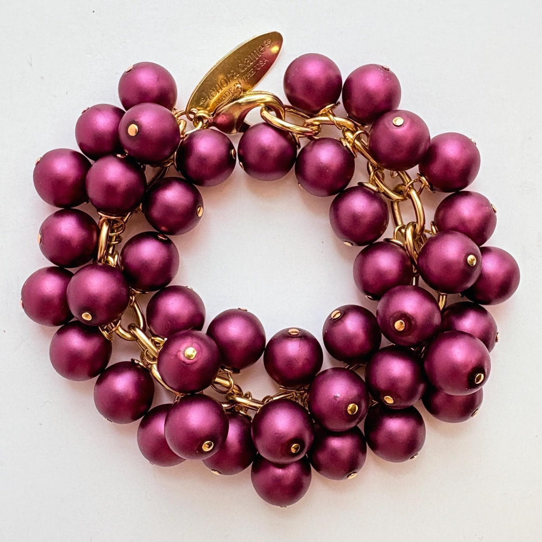 A gold chain bracelet with multiple strands of dark purple faux pearls. The bracelet has a small tag that reads &