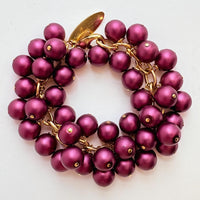 A gold chain bracelet with multiple strands of dark purple faux pearls. The bracelet has a small tag that reads &#39;Lenora Dame&#39; and &#39;Made in the USA.&#39;