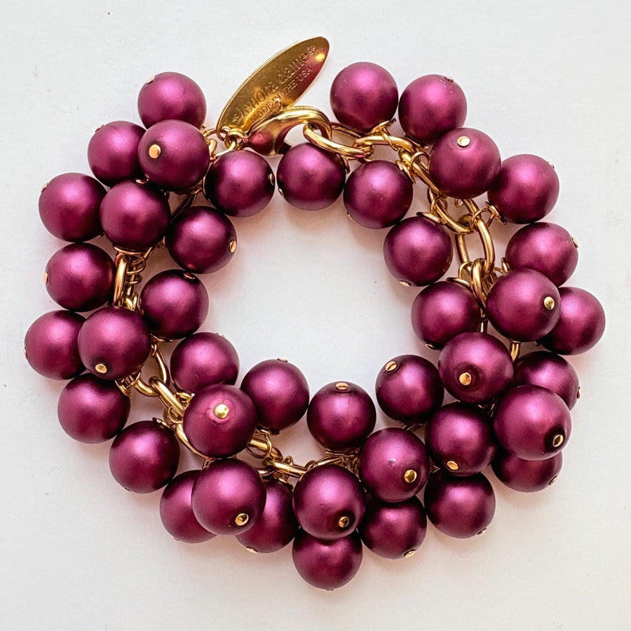A gold chain bracelet with multiple strands of dark purple faux pearls. The bracelet has a small tag that reads &#39;Lenora Dame&#39; and &#39;Made in the USA.&#39;