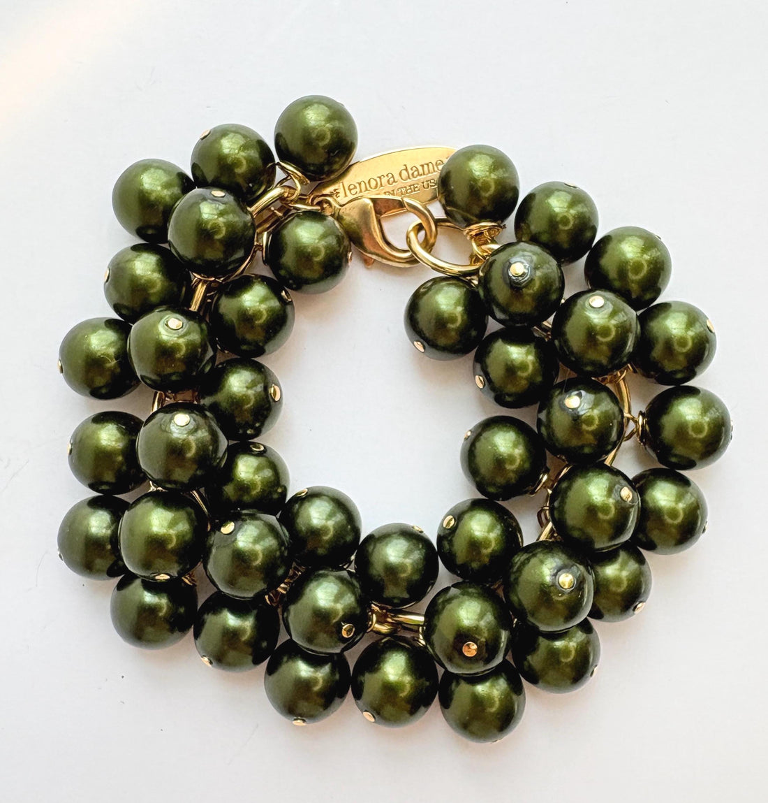 A gold chain bracelet with multiple strands of shiny hunter green faux pearls. The bracelet has a small tag that reads &