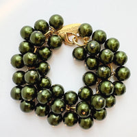 A gold chain bracelet with multiple strands of shiny hunter green faux pearls. The bracelet has a small tag that reads &#39;Lenora Dame&#39; and &#39;Made in the USA.&#39;