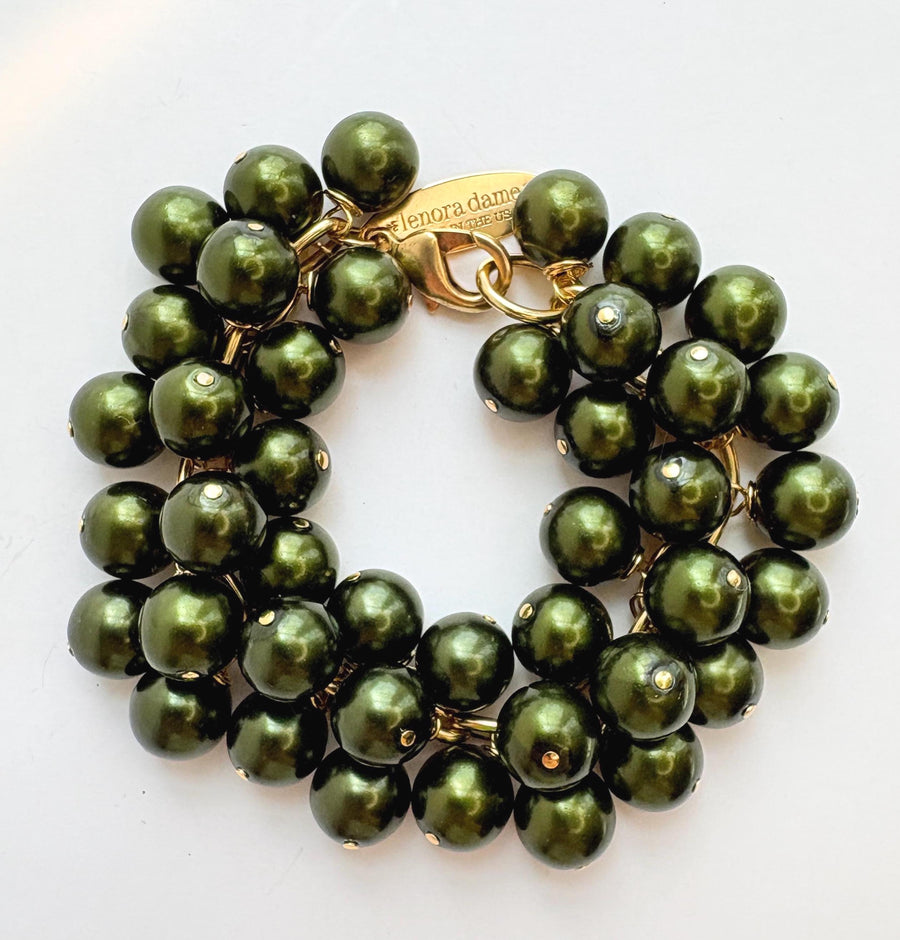 A gold chain bracelet with multiple strands of shiny hunter green faux pearls. The bracelet has a small tag that reads &#39;Lenora Dame&#39; and &#39;Made in the USA.&#39;