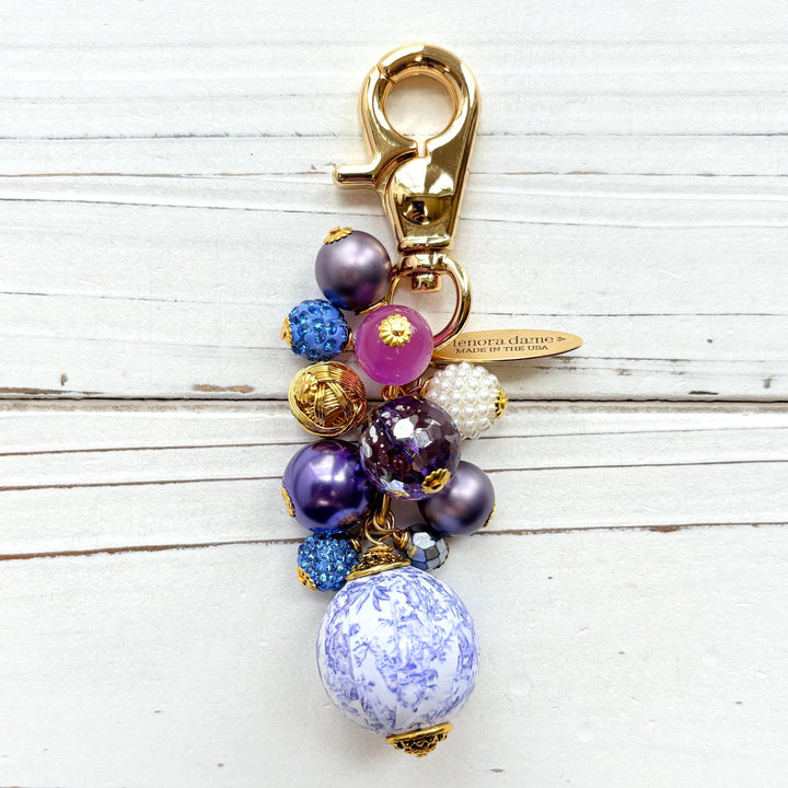 Beaded bag charm with a mix of bead styles in shades of purple, blue, gold and pearl. One larger round bead in a purple and white toile print hangs at the bottom and a gold trigger clasp on top of all of the beads. Brass oval hang tag near the trigger clasp that says &quot;Lenora Dame Handmade in USA&quot;. Lenora Dame bag charm.