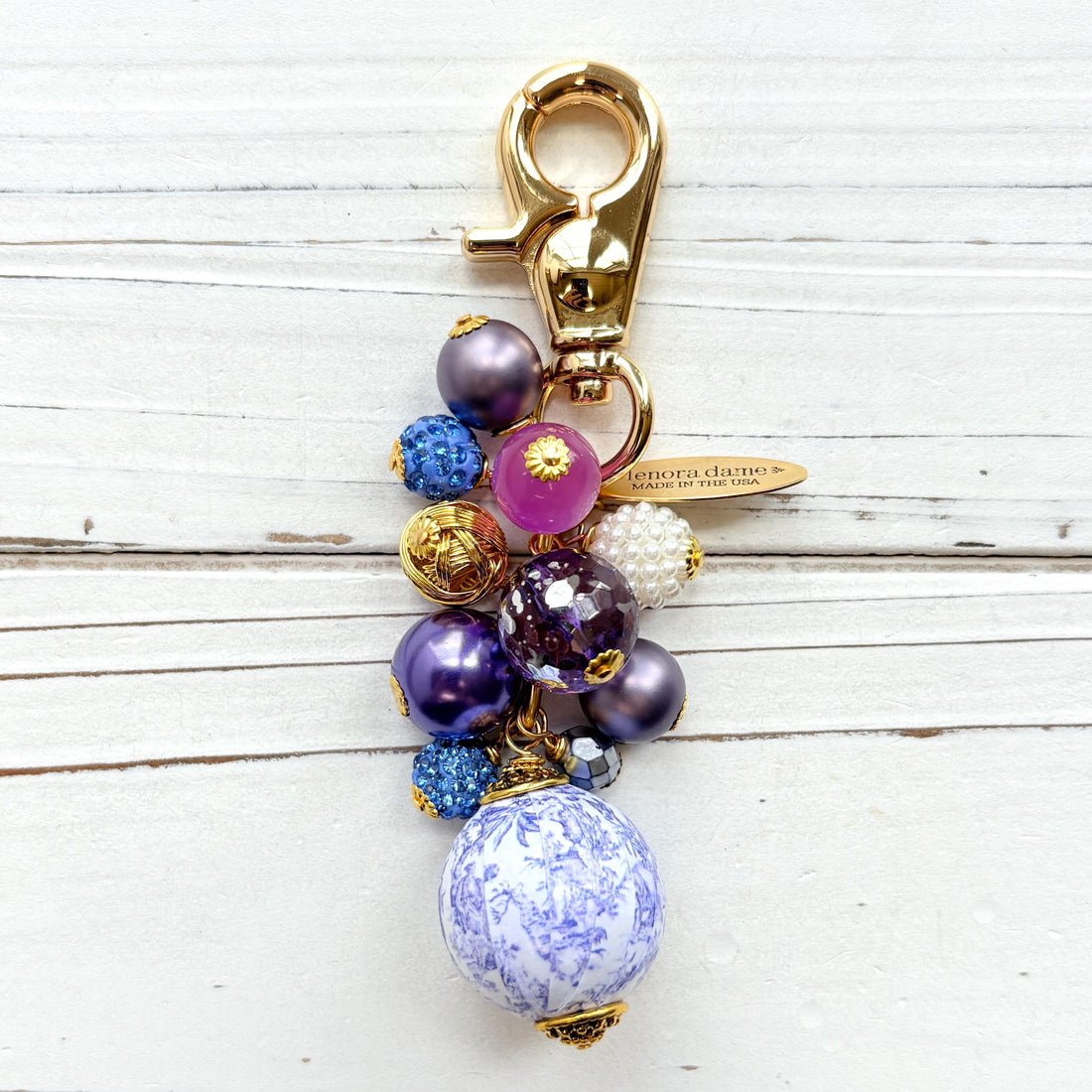 Beaded bag charm with a mix of bead styles in shades of purple, blue, gold and pearl. One larger round bead in a purple and white toile print hangs at the bottom and a gold trigger clasp on top of all of the beads. Brass oval hang tag near the trigger clasp that says &quot;Lenora Dame Handmade in USA&quot;. Lenora Dame bag charm.