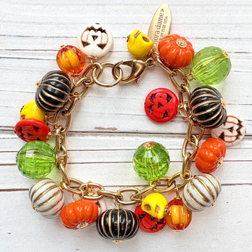 A gold chain bracelet with a variety of charms including green, orange, and white pumpkin charms, a skull charm, and a tag that reads Lenora Dame Made in the USA. Cute pumpkin charm bracelet.
