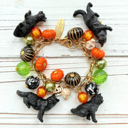 A gold chain bracelet with black cat charms, green and orange beads, and a white pumpkin charm. The bracelet has a tag that reads &#39;Lenora Dame&#39; and &#39;Made in USA&#39;.