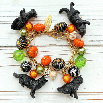 A gold chain bracelet with black cat charms, green and orange beads, and a white pumpkin charm. The bracelet has a tag that reads &#39;Lenora Dame&#39; and &#39;Made in USA&#39;.