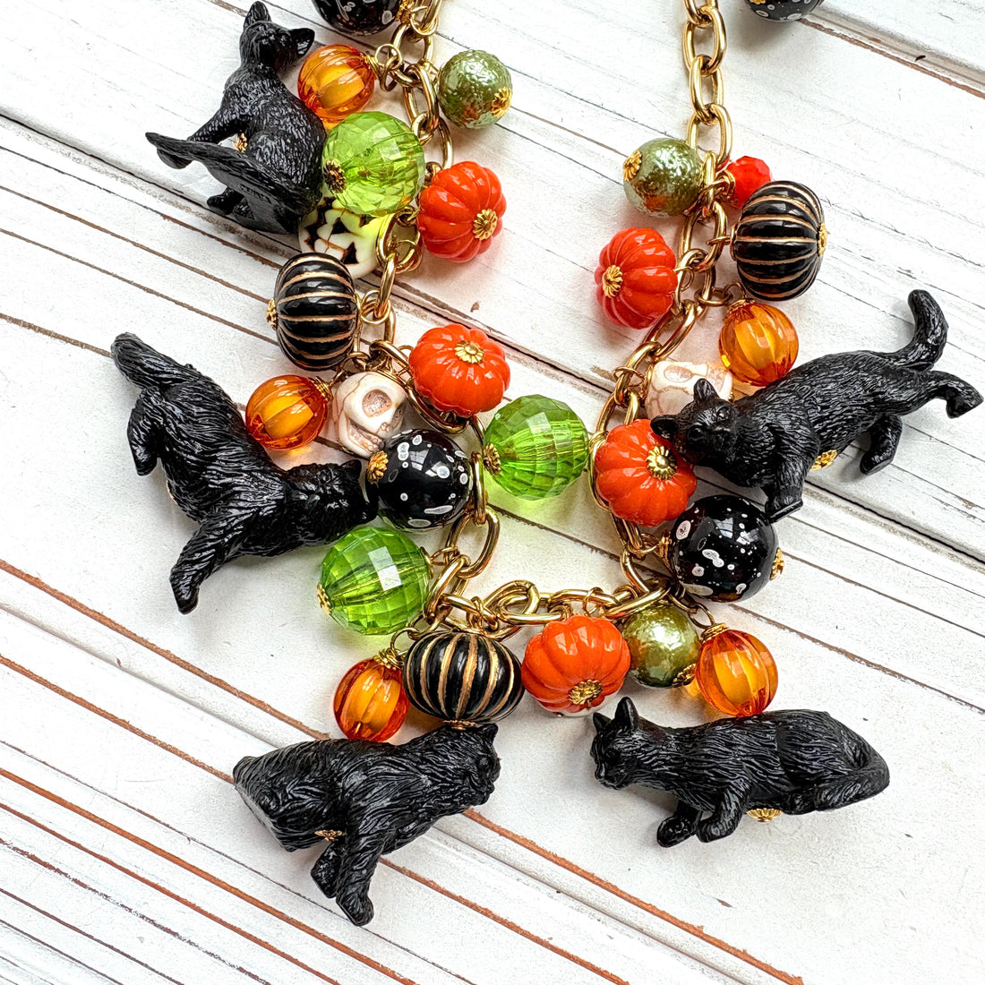 A gold chain necklace with black cat charms, orange, green, and white pumpkin charms, and black and white beads. The necklace is on a white wooden background. Lenora Dame cat necklace. Cute necklace for Halloween.