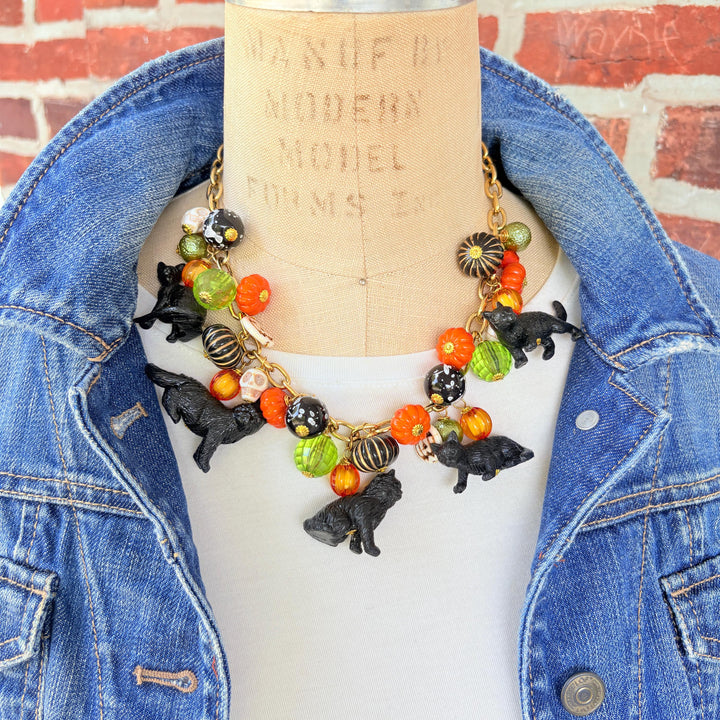 A gold chain necklace with black cat charms, orange, green, and white pumpkin charms, and black and white beads. The necklace is on a mannequin with a brick background.