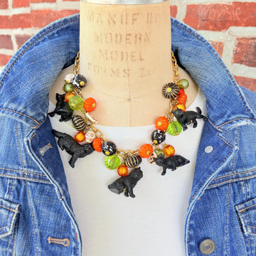 A gold chain necklace with black cat charms, orange, green, and white pumpkin charms, and black and white beads. The necklace is on a mannequin with a brick background.