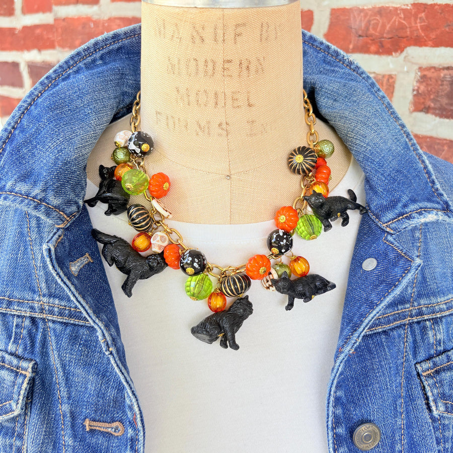 A gold chain necklace with black cat charms, orange, green, and white pumpkin charms, and black and white beads. The necklace is on a mannequin with a brick background.