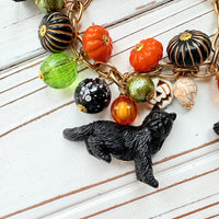 A gold chain bracelet with black cat charms, green and orange beads, and a white pumpkin charm. The bracelet has a tag that reads &#39;Lenora Dame&#39; and &#39;Made in USA&#39;.