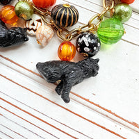 A gold chain bracelet with black cat charms, green and orange beads, and a white pumpkin charm. The bracelet has a tag that reads &#39;Lenora Dame&#39; and &#39;Made in USA&#39;.