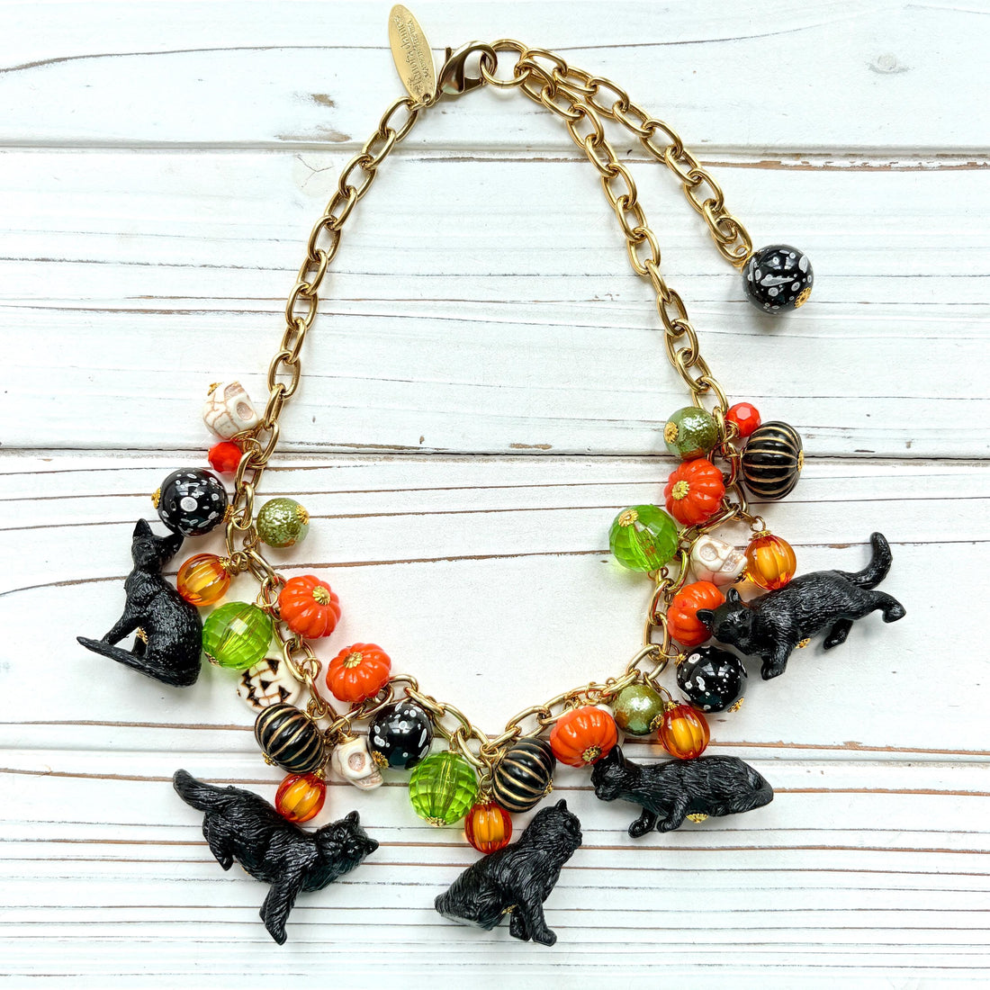 A gold chain necklace with black cat charms, orange, green, and white pumpkin charms, and black and white beads. The necklace is on a white wooden background. Lenora Dame cat necklace. Cute necklace for Halloween.