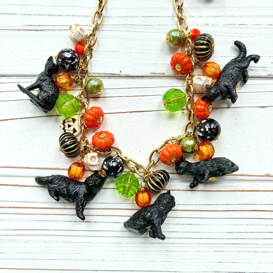 A gold chain necklace with black cat charms, orange, green, and white pumpkin charms, and black and white beads. The necklace is on a white wooden background. Lenora Dame cat necklace. Cute necklace for Halloween.