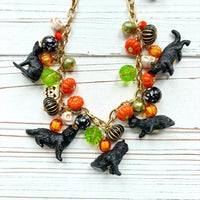 A gold chain necklace with black cat charms, orange, green, and white pumpkin charms, and black and white beads. The necklace is on a white wooden background. Lenora Dame cat necklace. Cute necklace for Halloween.