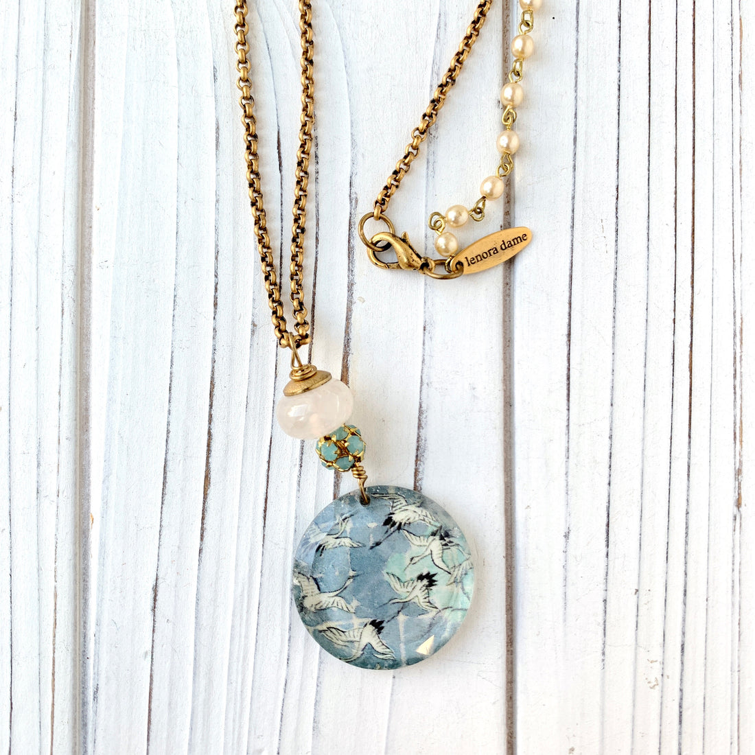 May include: A gold chain necklace with a round glass pendant featuring a blue and white pattern of flying birds. The pendant is attached to a white bead and a cluster of smaller blue beads. The necklace has a gold clasp and a small gold tag that reads &