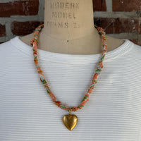 May include: A necklace with a peach-colored fabric cord, adorned with floral patterns in green, blue, and yellow. A large, gold-colored heart pendant hangs from the center, creating a vintage-inspired accessory.